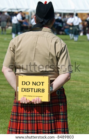 Judge at a Scottish games competition, wearing a kilt and black hat, holding a sign behind his back that says, \