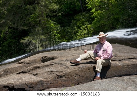 Senior man, wearing hat, sitting on rocks by flowing water in bright sunshine. He is staying active during retirement.