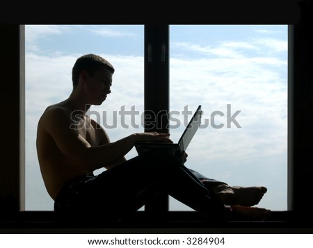 Silhouetted shirtless man working on a laptop sitting on the window-sill