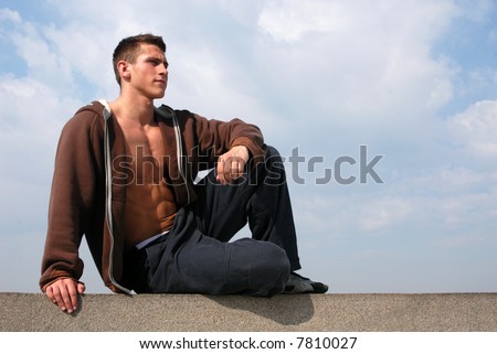 Young muscular man in a brown undone jacket sitting on the stone blocks