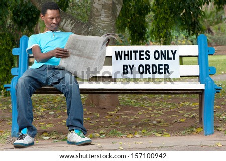 A non-white man sits on a bench in a park reserved for whites. This was commonplace during the apartheid years in South Africa.