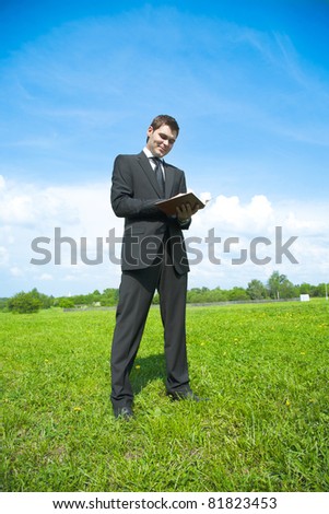 young adult businessman in a suit, standing in a meadow with notebook in hand and make it write