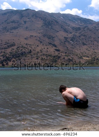 Backview of a young teenage man crouching and inspecting the water in a lake