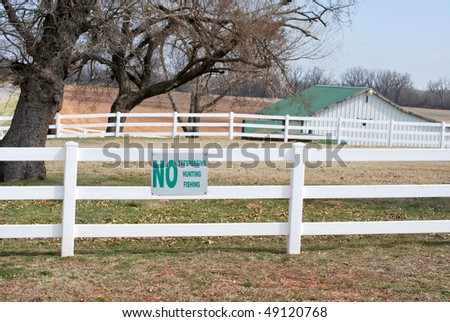 A photograph of a No Trespassing Hunting Fishing sign of a vinyl white fence with a barn in the background.