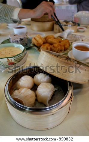 A table of dim sum (Chinese snack foods) in a Hong Kong restaurant, with a hand reaching in with chopsticks. A bamboo steamer with shrimp dumplings is in the foreground.