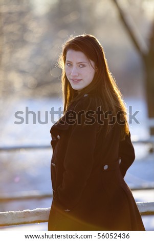 Backlit by the evening sun, the teenage girl turns her face to the camera,  in this winter portrait with snow covered woods in the background