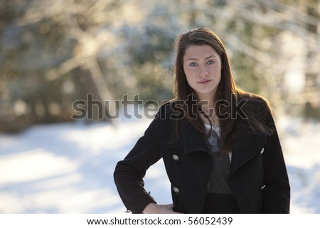 Backlit by the evening sun, the teenage is looking striaght into the camera,  in this winter portrait with snow covered woods in the background, where the sun is creating a nice halo behind the girl