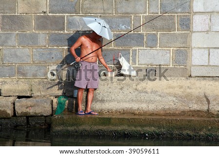 A Chinese fisherman with an umbrella hat and shorts on a riverbank