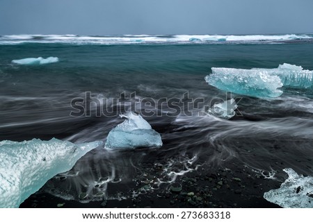Slow shutter photo showing wave movements around Ice blocks that get washed ashore at black sand beach in Iceland during sunset.