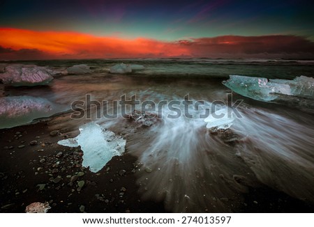 Slow shutter photo showing wave movements around Ice blocks that get washed ashore at black sand beach in Iceland during sunset.