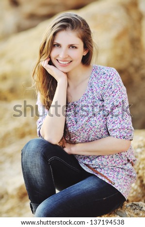 The attractive bright girl sits on the stone of a beach smiles