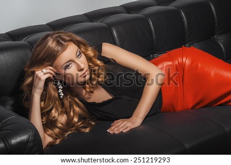 Beautiful blond in red leather skirt lying on the black leather divan. Long curly hairstyle. Jewelry. Vogue Style. Glamour makeup.