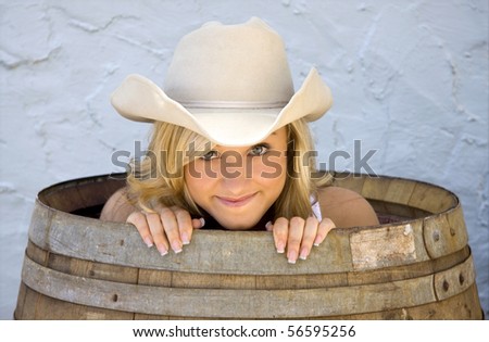 A beautiful young cowgirl peeks out of a barrel, looking like she has a secret.
