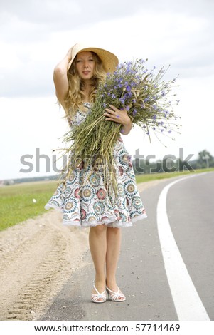 The woman in a dress costs on the brink of road with a bunch of flowers and holds with a hand a hat
