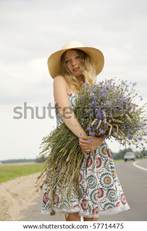 The woman in a hat with a bouquet of cornflowers costs on the brink of road near passing by the car