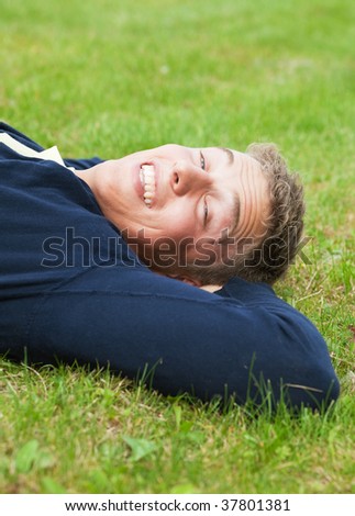 Young blond guy lying on grass, slit eyes because of bright sunlight