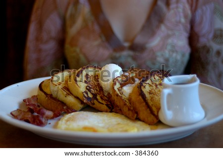 Plate with breakfast. French toast with butter, syrup, eggs and bacon. Woman in background.