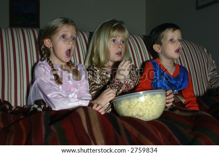 Movie Night:  Three children in their pajamas enjoy a movie in the dark while sharing a bowl of popcorn.