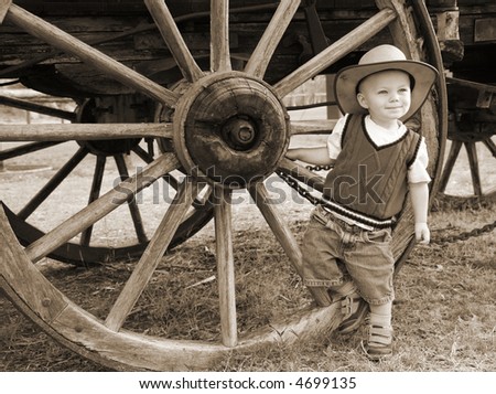 Sepia Farmboy:  A baby cowboy hangs out at the wheel of an old farm wagon in a cowboy hat and denim shorts.