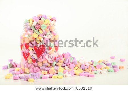 Candy conversation hearts spill over from a clear container decorated with red hearts against a white background with space for copy. Love/valentine\'s day concept.