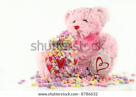 Soft and Sweet: A soft, pink teddy bear sitting next to an overflowing container of conversation hearts. Shallow DoF with focus on the container of candy. Valentine concept with space for copy.