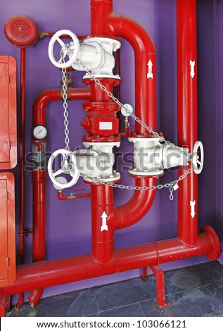 Red color fire fighting pipe system with an alarm bell hanging on a purple painted concrete wall.