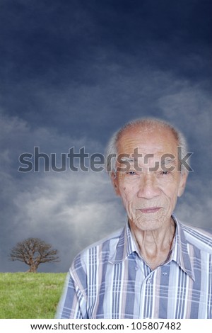 Portrait of an old wrinkly Chinese man in a park with a barren tree on top of a hill and a surreal cloudy sky in the background.