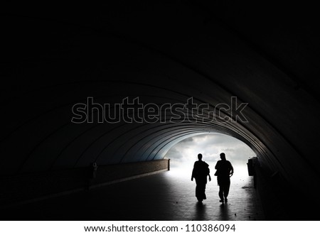 Silhouette of two woman walking into the light at the end of an underground pedestrian tunnel.