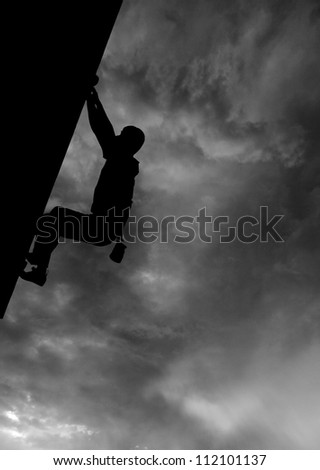 Silhouette of a rock climber hanging on to a cliff against a dramatic cloudy sky in monochrome.