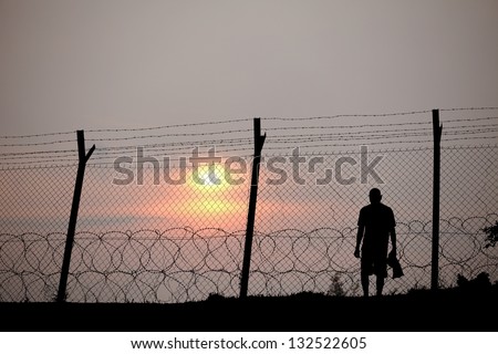 Silhouette Of A Prisoner Behind A Barbed Wire Fence In A Concentration Camp Against Fiery Setting Sun.
