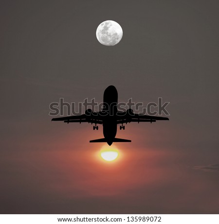 Silhouette of an airplane taking off against a fiery setting sun flying towards a full moon in the evening sky.
