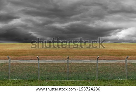 Security chain linked fence with barbed wire at the perimeter of a restricted area with dark stormy cloud in the horizon.