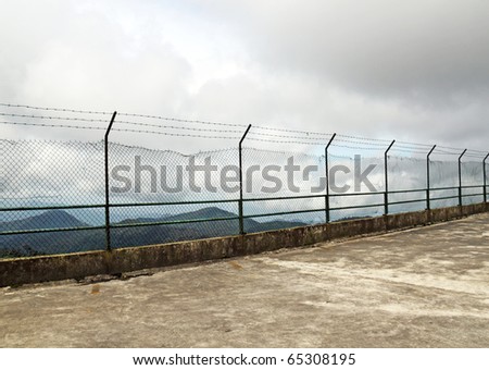 An image of a barbed wire high security fence on the ledge of a cliff with a view of a mountainous range.
