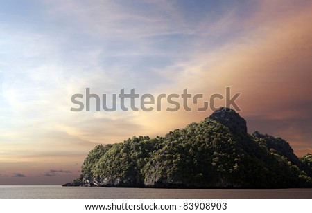 A remote rural rocky outcrop island covered with green vegetation in a calm sea against a surreal candy color sunset.