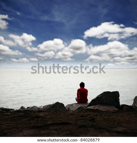 A lonely man in deep thought while sitting on a rocky bank facing the sea with a surreal cloudy blue sky.