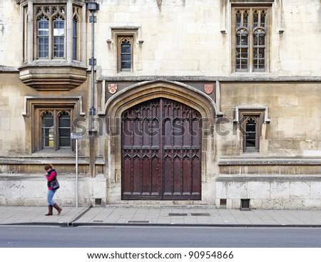 The facade of a medieval brownstone building with a large ornate timber door in Oxford city, England, with a young lady student in motion across it.