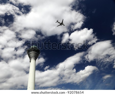 An airplane flying overhead across an airport control tower against a blue cloudy sky.
