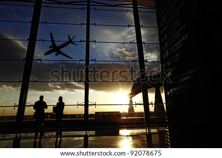 Silhouette of airline passengers in an airport lounge at the wide observation window watching an airplane flying of against a surreal sunset in the horizon with direction signage in the foreground.