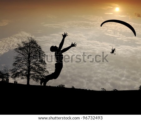 Silhouette of a young boy jumping in excitement on a field at the sight of a paraglider gliding across a fiery sunset sky.