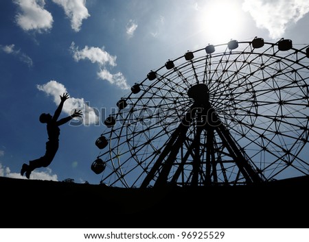 Silhouette of a young boy jumping in excitement at a ferris wheel in an amusement park on a blue cloudy day.