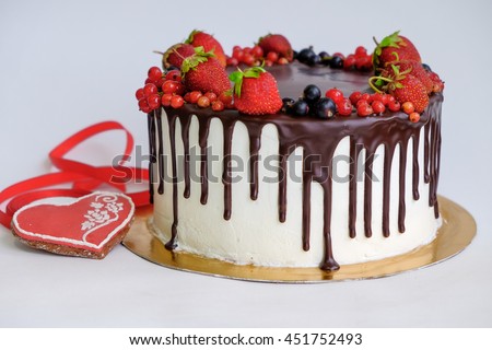 birthday cake decorated by chocolate and berry, strawberry on top near red gingerbread like heart shape and red ribbon on white background/different focus and angle