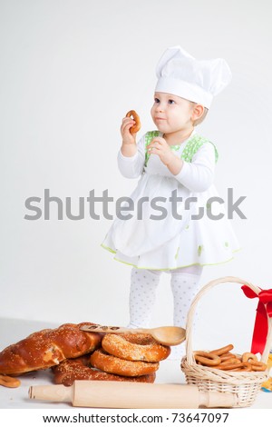 Little baby girl in the cook costume standing near bread rolls and bagels. She is eating bagel.