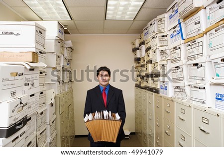 A young businessman is standing in a file room holding files. Cabinets and storage boxes line the sides. Horizontal shot.
