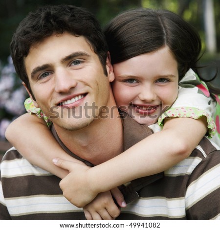 Young girl wraps her arms around a young man from behind. They are both smiling towards the camera. Square shot.