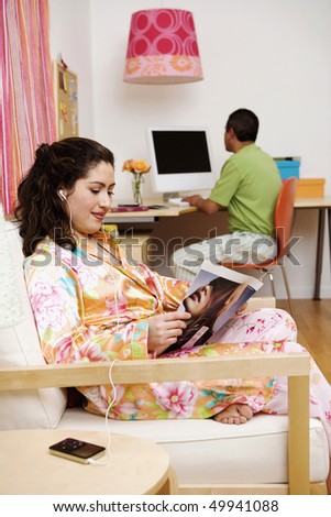 A woman reads a magazine and listens to music on a futon while her husband works on a computer in the background. Vertical shot.