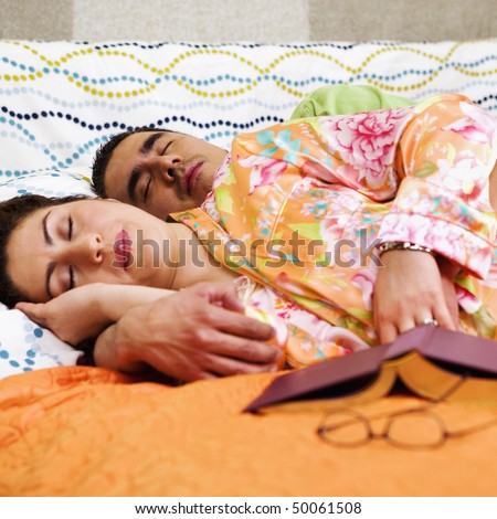 A man and woman sleep on a bed with a book and eye glasses in the foreground. Square format.