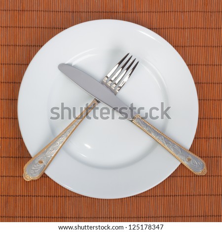 White plate, knife and fork crossed each other on the brown tablecloth from bamboo