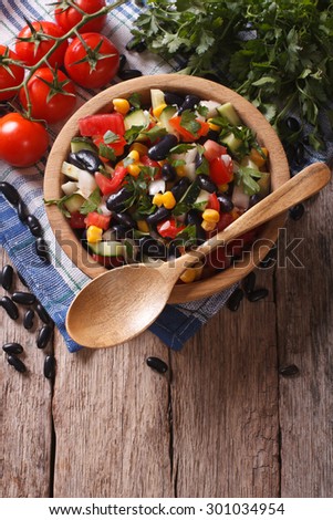 Mexican vegetable salad with black beans in a wooden bowl, close-up and ingredients on the table. vertical top view