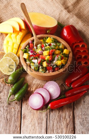 Mexican salsa with mango, cilantro, onions and hot peppers close up in a wooden bowl and ingredients on the table. vertical