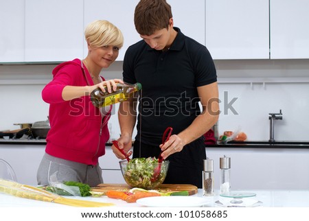 Portrait of a lovely couple making a salad in their kitchen. blond girl and dark haired guy. he mixes salad and she pours olive oil. kitchen is white and filled with light. couple is young and fit